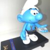 Blauwe smurf sculptuur met Louis Vuitton logo, heuptasje en dollars op een luxe houten plateau, pop-art assemblage kunst.
