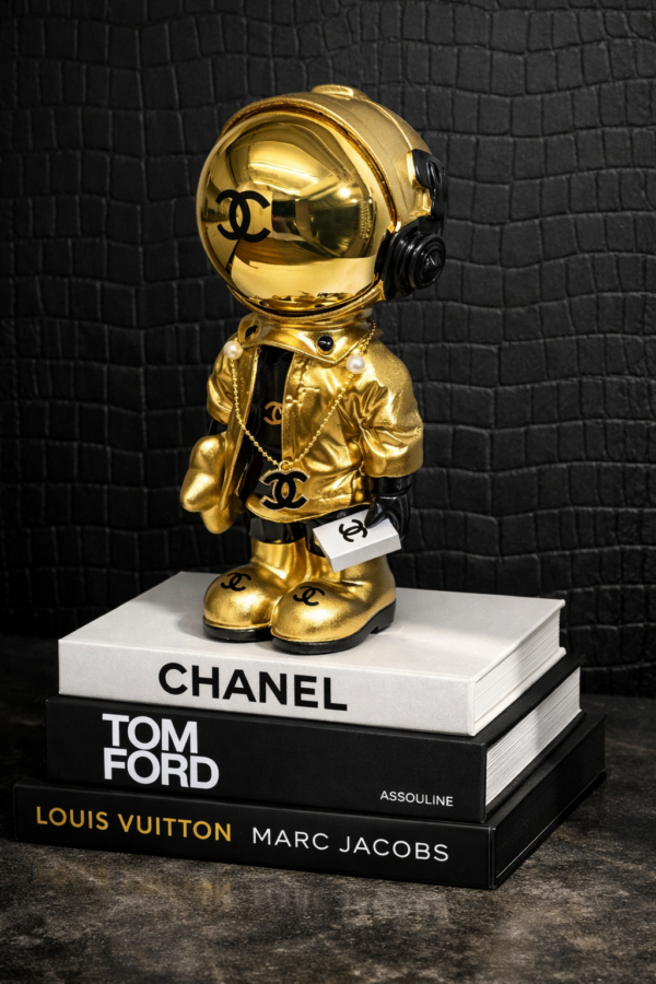 Astro Coco Gold Gouden Chanel‑astronautbeeldje op stapel designboeken met matte zwarte croco‑achtergrond