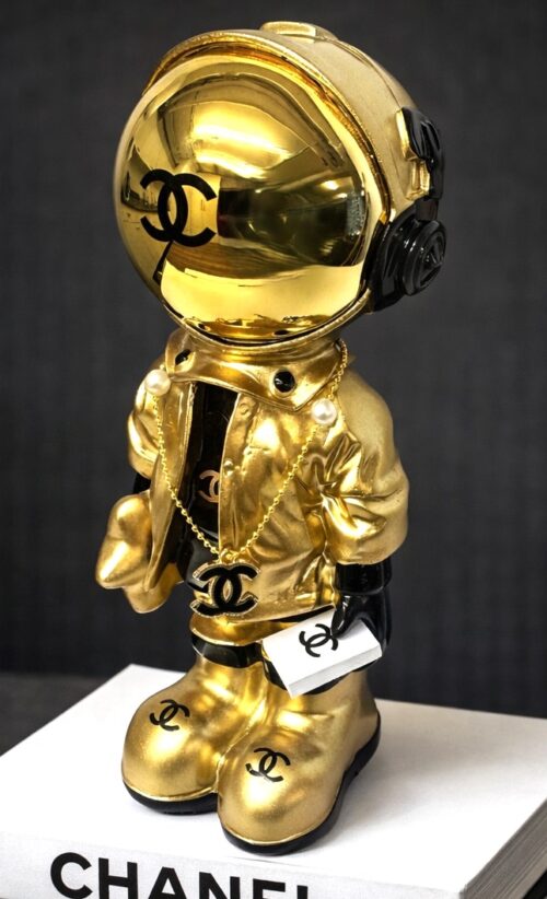 Astro Coco Gold Gouden Chanel‑astronautbeeldje op stapel designboeken met matte zwarte croco‑achtergrond